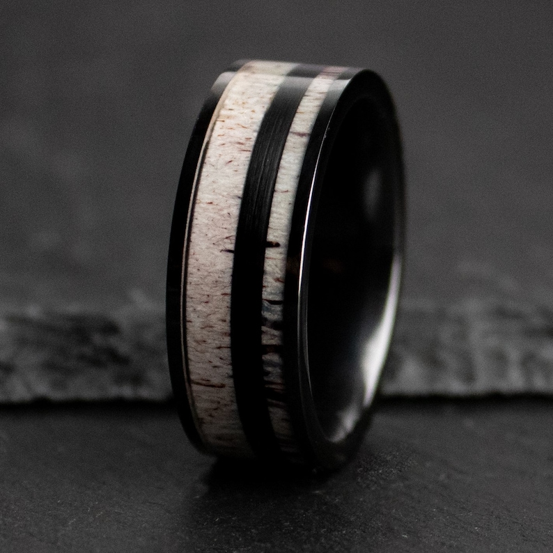 Puede incluir: Un anillo negro con una banda central de material claro, posiblemente asta o hueso, flanqueada por dos bandas negras. El anillo tiene una superficie pulida y reflectante, y el dise&ntilde;o es sim&eacute;trico.