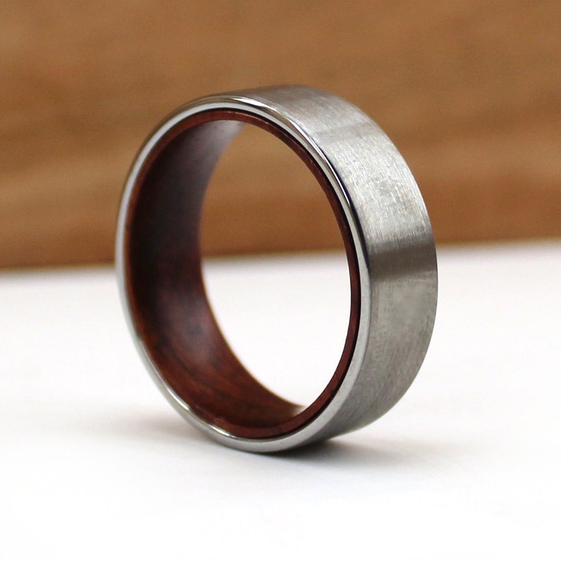 May include: A close-up of a silver ring with a brushed metal finish. The ring has a dark reddish-brown wooden interior. The ring is set against a white and brown background.