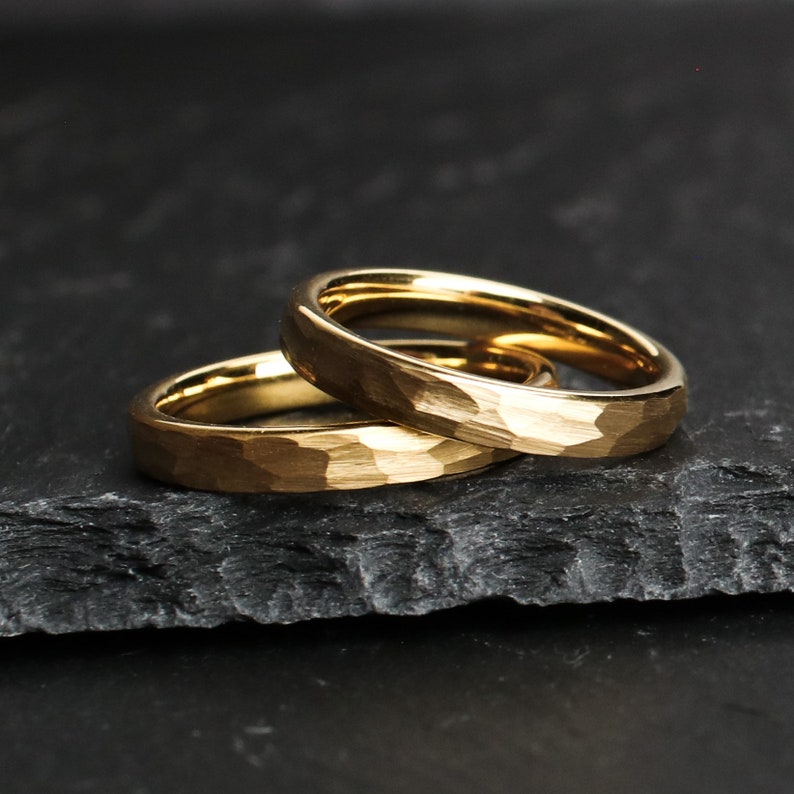 May include: Two gold-colored rings with a hammered texture are stacked on top of each other. The rings are set against a dark, textured background. The rings have a simple, elegant design.