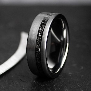 May include: A close-up of a dark grey ring with a brushed metal finish. The ring features a black, textured inlay that runs around the centre. The ring is a wedding band or fashion accessory.