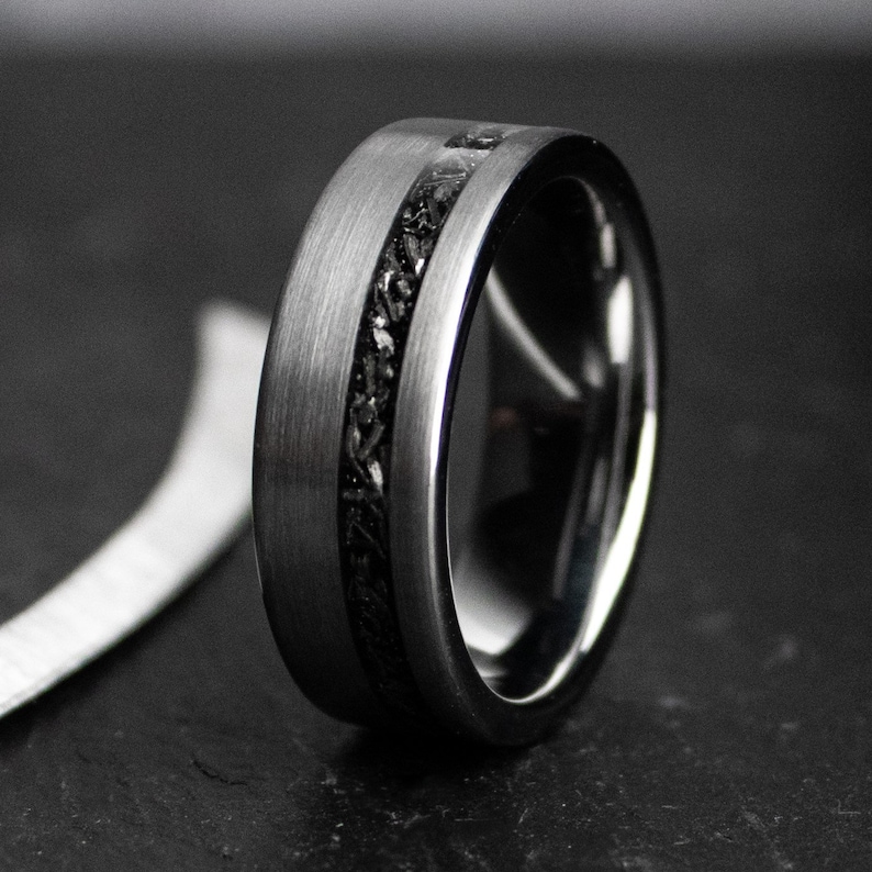 May include: A close-up of a dark grey ring with a brushed metal finish. The ring features a black, textured inlay that runs around the centre. The ring is a wedding band or fashion accessory.
