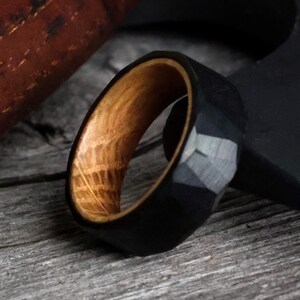 May include: A black faceted ring with a light brown wooden interior. The ring has a geometric design and is set against a wooden surface. The ring's interior wood grain is visible.
