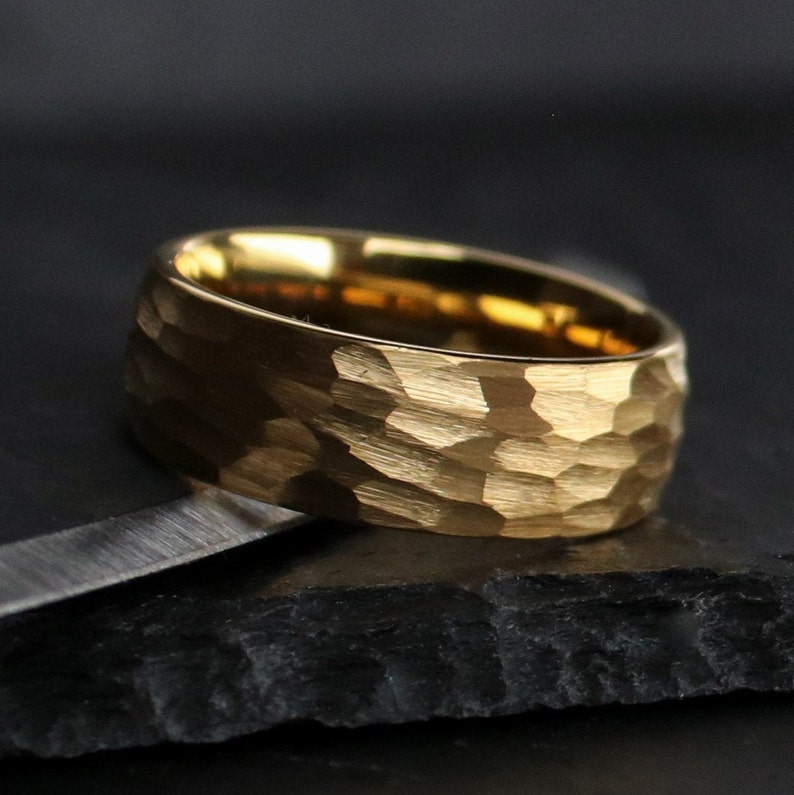 May include: A gold-colored ring with a textured, hammered finish. The ring has a wide band and a matte appearance. The ring is displayed on a dark, textured surface.