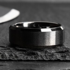 May include: A close-up of a black ring with a brushed metal finish. The ring has a wide band and a beveled edge. The ring is on a dark, textured surface.