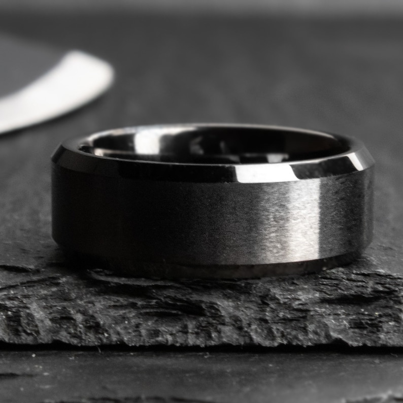 May include: A close-up of a black ring with a brushed metal finish. The ring has a wide band and a beveled edge. The ring is on a dark, textured surface.