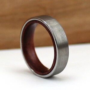 May include: A close-up of a ring with a brushed silver exterior and a dark reddish-brown wood interior. The ring has a smooth, polished finish and a simple, elegant design. The ring is a wedding band or fashion accessory.