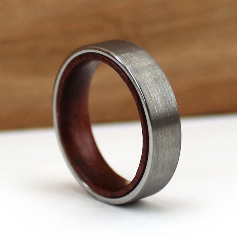 May include: A close-up of a ring with a brushed silver exterior and a dark reddish-brown wood interior. The ring has a smooth, polished finish and a simple, elegant design. The ring is a wedding band or fashion accessory.