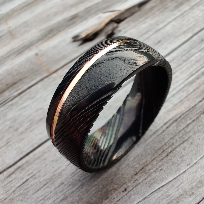 May include: A black Damascus steel ring with a gold inlay. The ring has a textured surface and a polished gold stripe running along the center. The ring is displayed on a light wood surface.