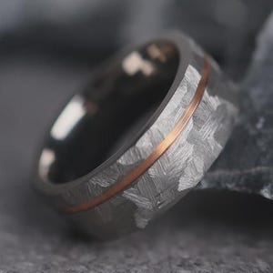 May include: Close-up of a textured, gray ring with a thin, copper-colored band. The ring has a woven pattern and a polished, metallic appearance. The ring is a piece of jewelry.