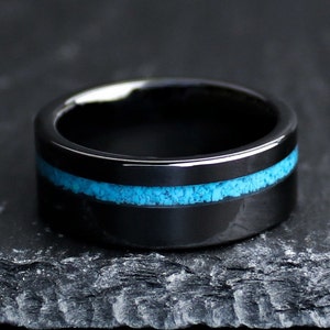 May include: A black ring with a bright turquoise inlay. The ring is made of a dark, glossy material and sits on a dark, textured surface. The turquoise inlay is a band of small, irregular pieces.
