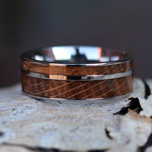 Puede incluir: Primer plano de un anillo con dise&ntilde;o de barril de madera. El anillo tiene una banda plateada en la parte superior e inferior, con una franja plateada en el medio del dise&ntilde;o de barril de madera. El anillo est&aacute; sobre una superficie clara.