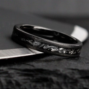 May include: A black ring with a textured band and a silver inlay design. The ring is set against a dark background, highlighting its details. The ring's design is intricate and eye-catching, suitable for everyday wear or special occasions.