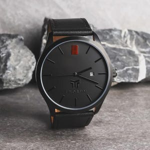 May include: A black wristwatch with a leather band. The watch face is black with a red rectangular marker at the 12 o'clock position. The watch has the brand name "THORUM" printed on the face. The watch is set against a gray stone background.