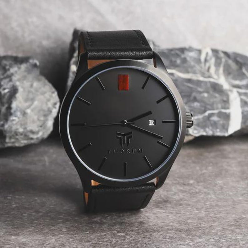 May include: A black wristwatch with a leather band. The watch face is black with a red rectangular marker at the 12 o'clock position. The watch has the brand name "THORUM" printed on the face. The watch is set against a gray stone background.