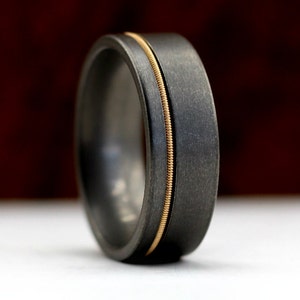 May include: A dark grey ring with a gold-coloured inlay. The ring has a brushed metal finish and a simple, modern design. The gold inlay runs vertically through the centre of the ring.