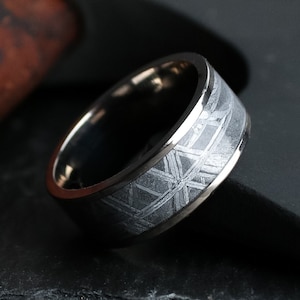 Op de afbeelding: Close-up van een zilveren ring met een patroonband. De ring heeft een donkergrijs, geometrisch ontwerp en een gepolijste zilveren afwerking. De ring staat tegen een donkere achtergrond.