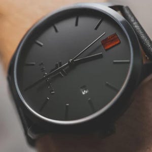 May include: Close-up of a black wristwatch with a black leather band. The watch face is black with minimalist hour markers and hands. A small, rectangular orange accent is at the 3 o'clock position. The watch is a modern, minimalist design.