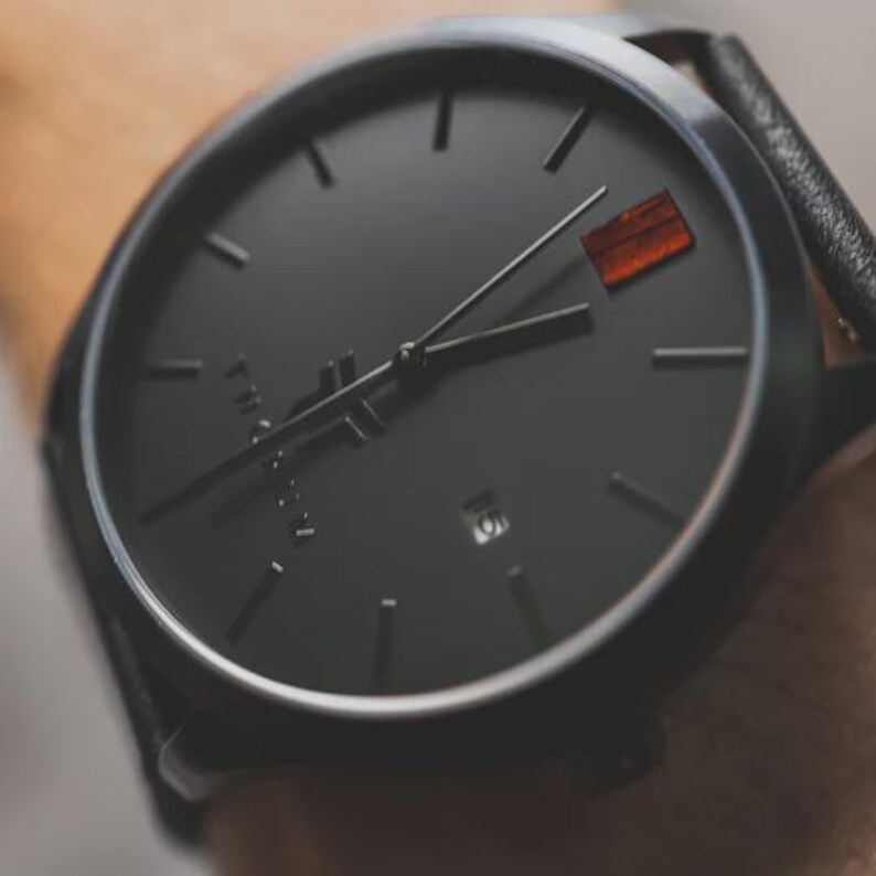 May include: Close-up of a black wristwatch with a black leather band. The watch face is black with minimalist hour markers and hands. A small, rectangular orange accent is at the 3 o'clock position. The watch is a modern, minimalist design.