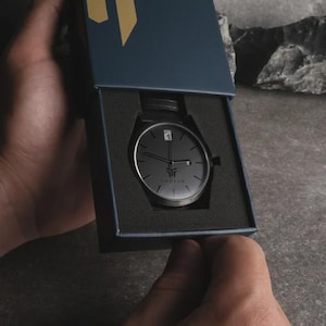 May include: A black wristwatch with a minimalist design, presented in a dark blue box. The watch features a black face with silver hands and a leather strap. The brand name "THOKUM" is visible on the watch face.