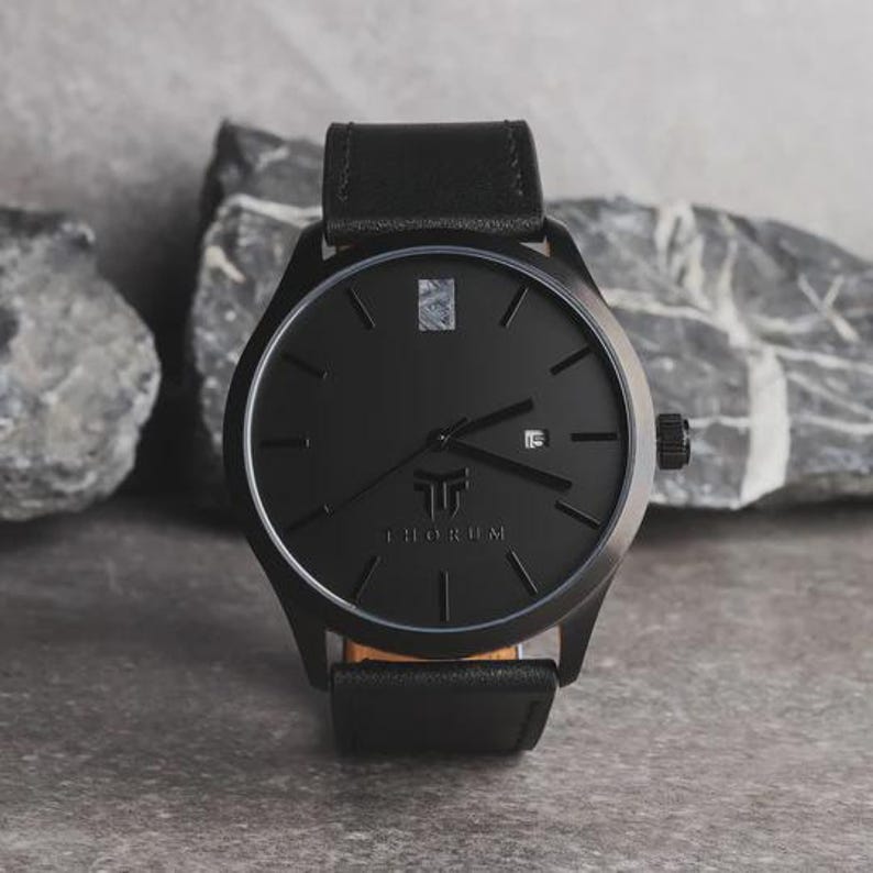 May include: A black wristwatch with a leather band. The watch face is black with silver accents and the brand name "THORUM" is displayed. The watch has a date display at the 3 o'clock position and a black crown.