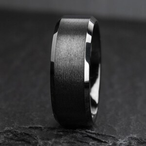 May include: A close-up of a black ring with a brushed metal finish. The ring has a flat top and beveled edges with a polished, reflective surface. The ring is a dark grey colour.