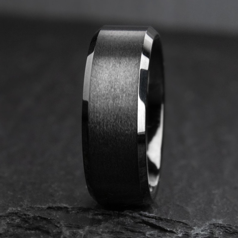 May include: A close-up of a black ring with a brushed metal finish. The ring has a flat top and beveled edges with a polished, reflective surface. The ring is a dark grey colour.