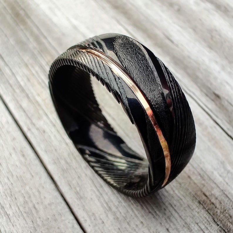 May include: A black ring with a textured surface and a thin, copper-colored stripe. The ring has a unique pattern and is set against a light wood grain background. The ring's design is eye-catching and modern.