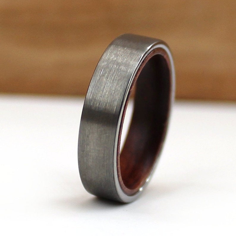 May include: A close-up of a silver ring with a brushed metal finish. The ring has a dark wood inlay and a thin silver band on the inside. The ring is approximately 0.6 cm wide.
