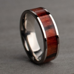 May include: A close-up of a ring with a dark metal band and a central inlay of reddish-brown wood. The wood grain is visible, and the ring has a polished, reflective surface. The ring is a piece of jewelry.