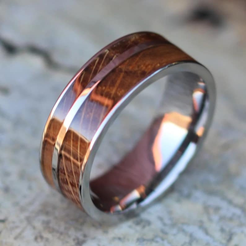 Puede incluir: Primer plano de un anillo con una banda de acero inoxidable y una incrustaci&oacute;n de madera. La madera es de color marr&oacute;n oscuro con vetas visibles. La superficie pulida del anillo refleja la luz.
