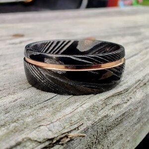 May include: A close-up of a black ring with a patterned design and a thin, rose gold band. The ring is set on a weathered wooden surface, showcasing its intricate details and contrasting colors.