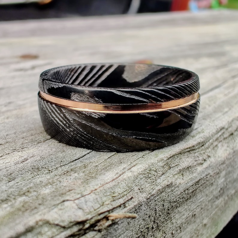 May include: A close-up of a black ring with a patterned design and a thin, rose gold band. The ring is set on a weathered wooden surface, showcasing its intricate details and contrasting colors.