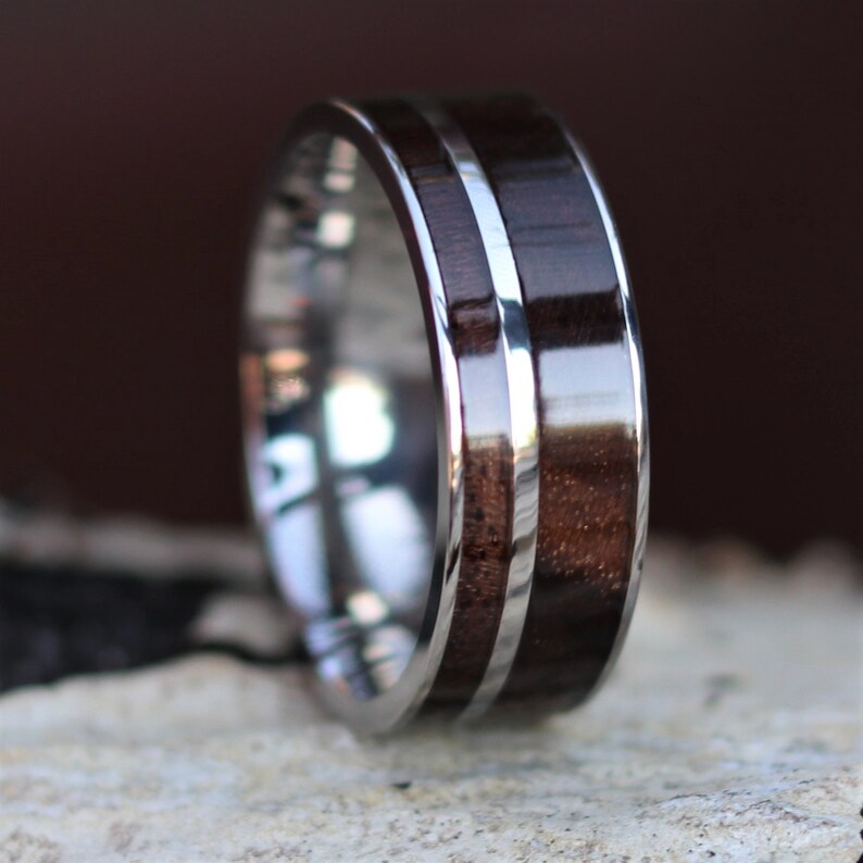 May include: A close-up of a ring with a dark brown wood inlay and a silver band. The wood grain is visible, and the silver band has a polished, reflective surface. The ring is set against a neutral background.
