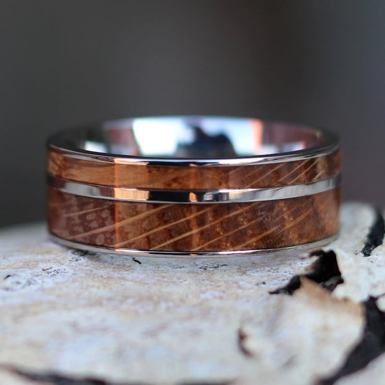 Puede incluir: Primer plano de un anillo con dise&ntilde;o de barril de madera. El anillo tiene una banda plateada en la parte superior e inferior, con una franja plateada en el medio del barril de madera. La madera es marr&oacute;n con l&iacute;neas diagonales.