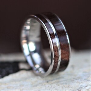 May include: A close-up of a silver ring with a dark brown wood inlay. The ring has a polished, metallic band with a central strip of dark wood. The wood grain is visible, and the ring appears to be well-crafted.