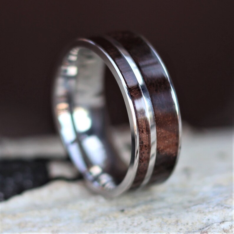 May include: A close-up of a silver ring with a dark brown wood inlay. The ring has a polished, metallic band with a central strip of dark wood. The wood grain is visible, and the ring appears to be well-crafted.
