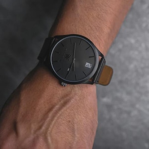May include: A black wristwatch with a black face and a leather band is shown. The watch has a minimalist design with a date window at the 3 o'clock position. The watch is on a wrist, and the background is a dark gray.