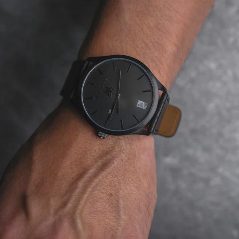 May include: A black wristwatch with a black face and a leather band is shown. The watch has a minimalist design with a date window at the 3 o'clock position. The watch is on a wrist, and the background is a dark gray.