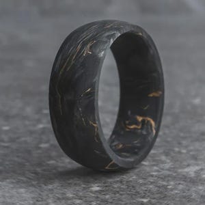 May include: A black ring with gold marbling. The ring is a solid band with a smooth, rounded exterior. The gold veining creates a unique pattern throughout the ring's surface. The ring is set against a gray background.