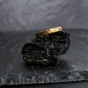 May include: A gold-colored ring with a hammered texture rests on a black, rough-hewn stone. The ring's surface reflects light, showcasing its textured design. The background is a blurred gray, providing contrast to the ring and stone.