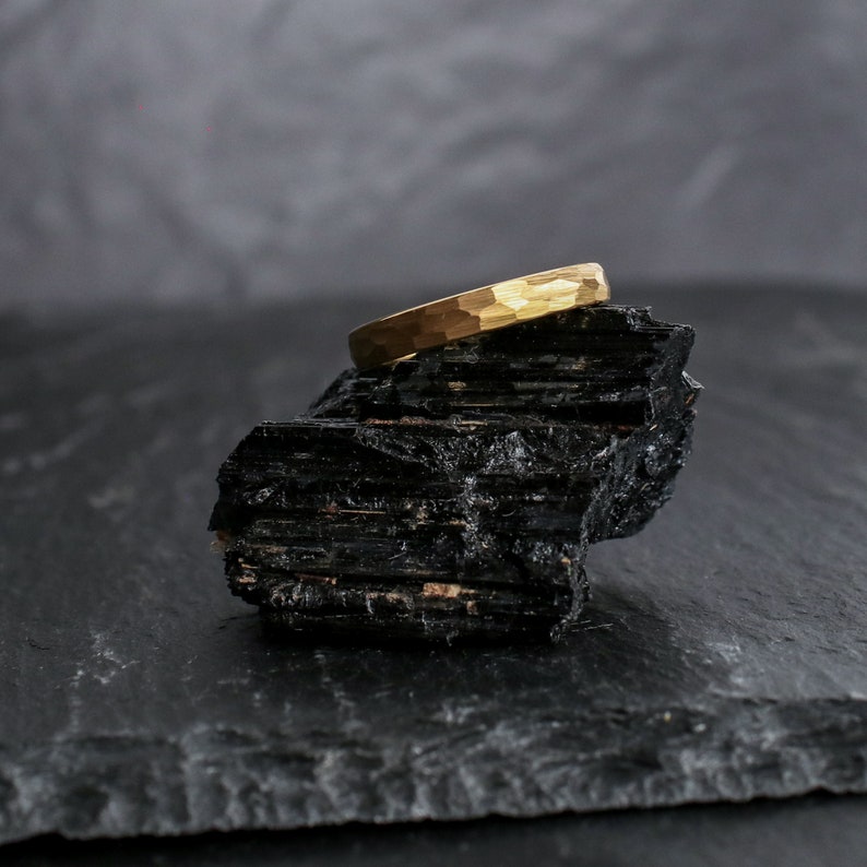 May include: A gold-colored ring with a hammered texture rests on a black, rough-hewn stone. The ring's surface reflects light, showcasing its textured design. The background is a blurred gray, providing contrast to the ring and stone.