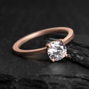 May include: A rose gold ring with a large, clear, round-cut gemstone. The ring has a thin band and a four-prong setting. The gemstone sparkles, reflecting light. The ring is displayed on a dark, textured surface.