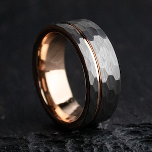 May include: A close-up of a tungsten ring with a hammered finish. The ring features a dark gray exterior with a thin rose gold band in the center and a rose gold interior. The ring is set against a dark background.