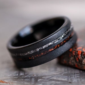 May include: A black ring with a textured band. The ring features a silver inlay and a band of orange and red speckled material. The ring is shown close up, with a blurred background.