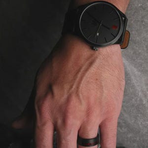 May include: A black wristwatch with a black dial and a brown leather strap. The watch is on a wrist, and a black ring with a red stripe is on a finger.
