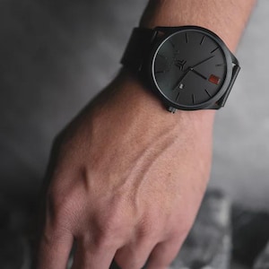 May include: A black wristwatch with a round face and a black leather band. The watch has a minimalist design with black hands and markers. A small red accent is visible on the watch face. The watch is on a wrist.
