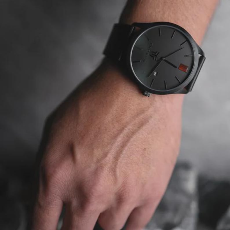 May include: A black wristwatch with a round face and a black leather band. The watch has a minimalist design with black hands and markers. A small red accent is visible on the watch face. The watch is on a wrist.