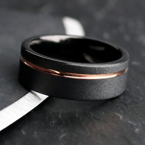 May include: A black ring with a textured band and a thin rose gold stripe. The ring has a polished interior and is set against a dark background. A modern piece of jewelry.