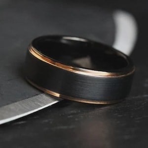 May include: Close-up of a black ring with a brushed metal finish and a thin gold-colored band around the edges. The ring is set against a dark background, with a metallic object in the foreground.