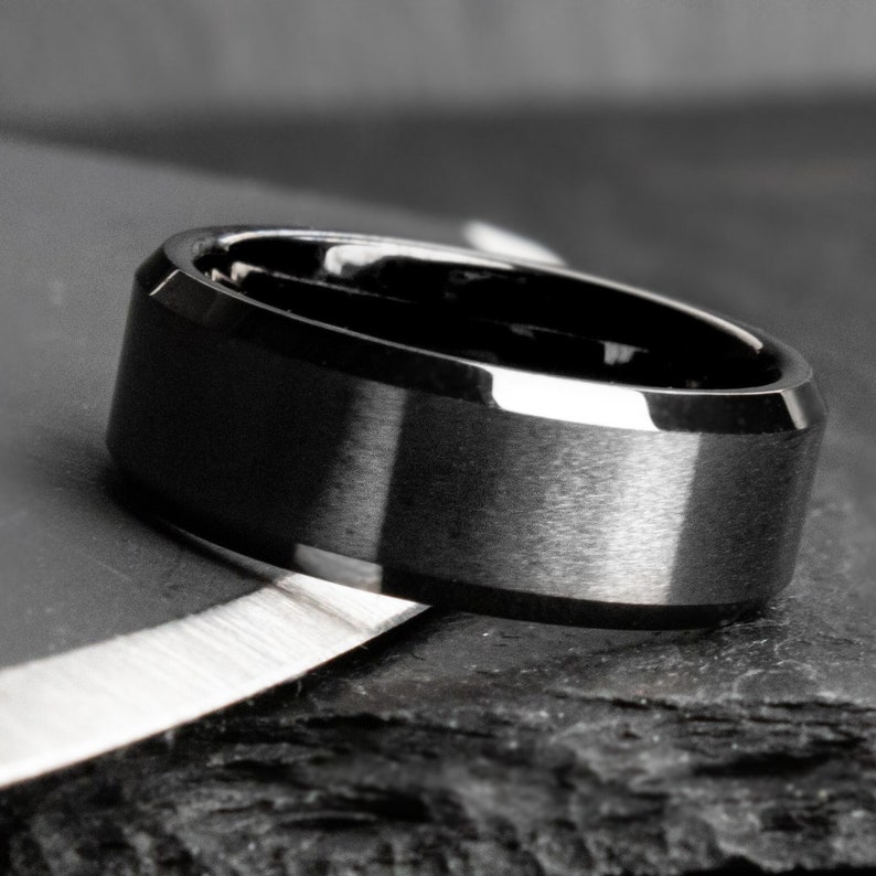May include: A black ring with a brushed metal finish. The ring has a flat top and beveled edges. The ring is shown on a dark, textured surface, with a silver object in the foreground.